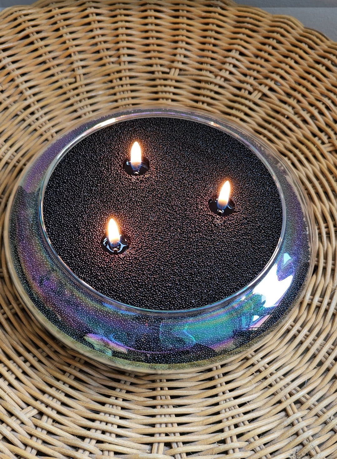 Three-wick black candle sand displayed in a round iridescent glass bowl on a woven rattan table mat.