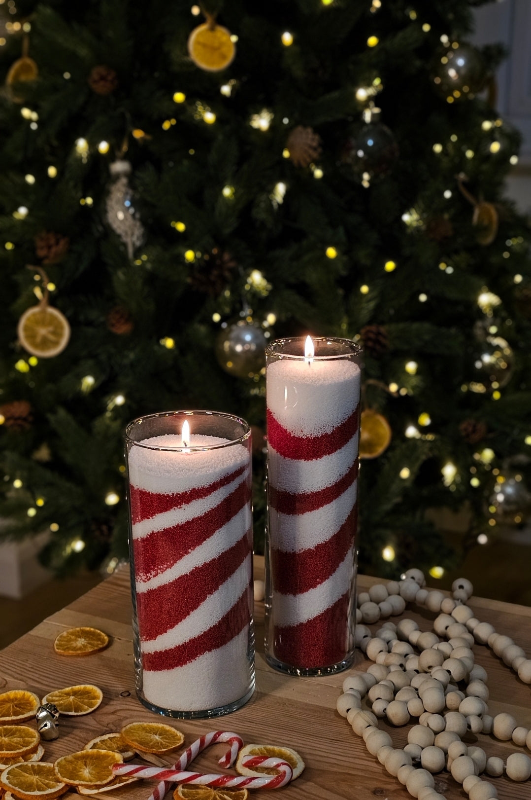 Red and white candy cane candle sand layered in two glass cylinders beside a Christmas tree.