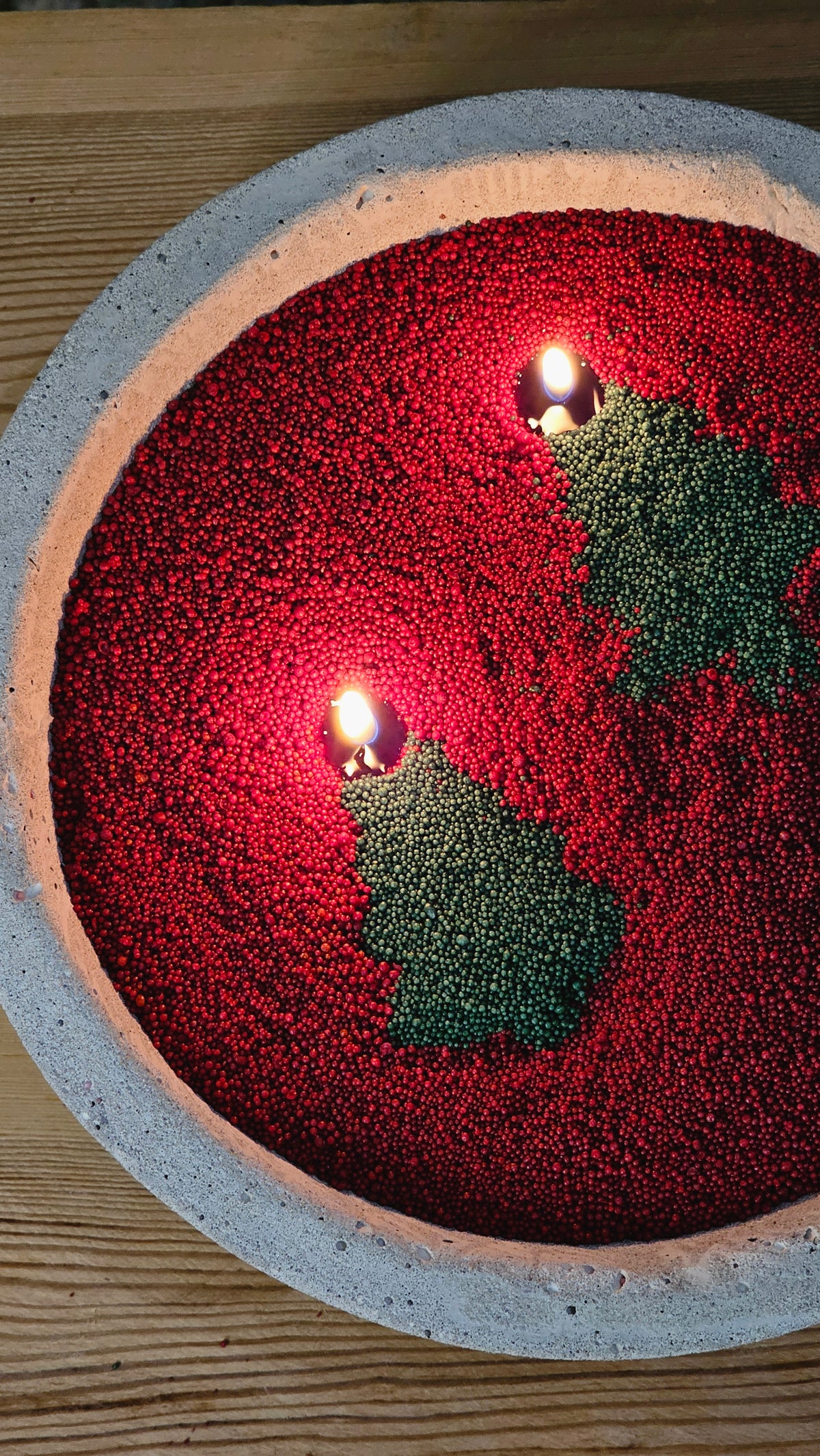 Red and green Christmas candle art made from coloured wax pearls with two lit wicks in a bowl.