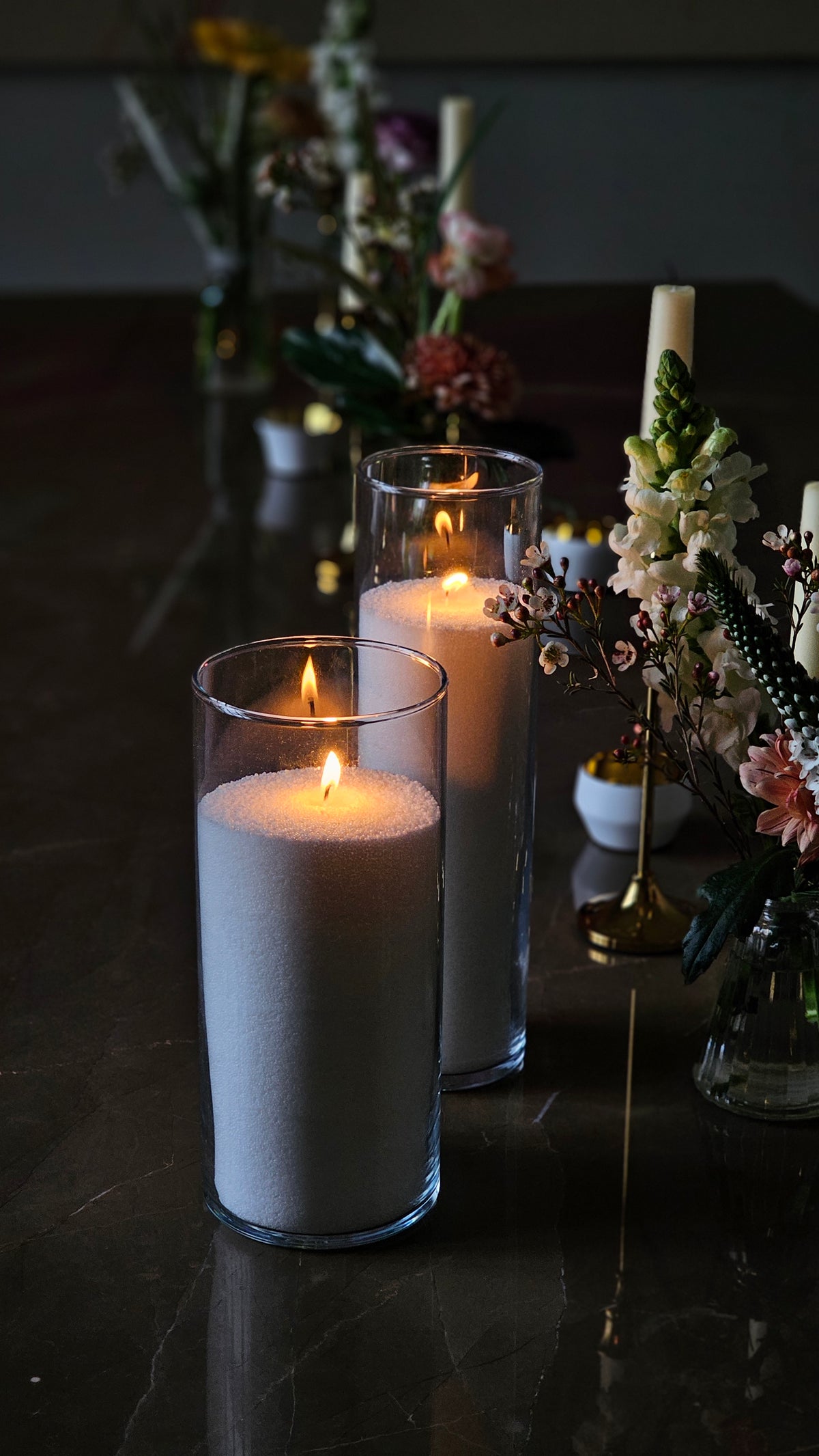 Two tall glass cylinder candles filled with white wax pearls and multiple wicks, styled with flowers as a table centrepiece.