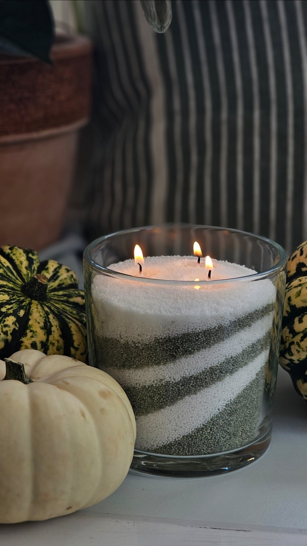 Green and white layered candle sand in a glass jar with two wicks, styled with pumpkins for autumn.