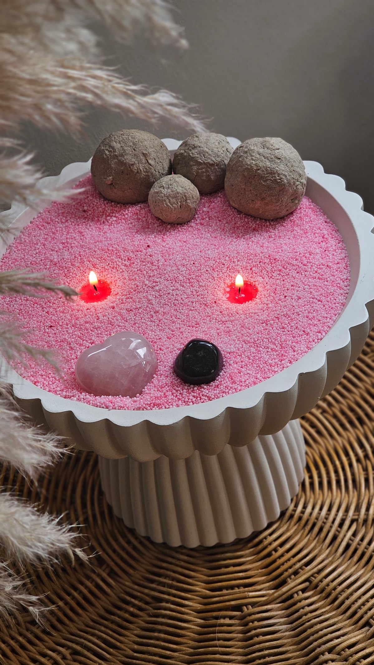 Pink candle sand in a white pedestal bowl with two lit wicks, polished crystals and decorative seed pods.