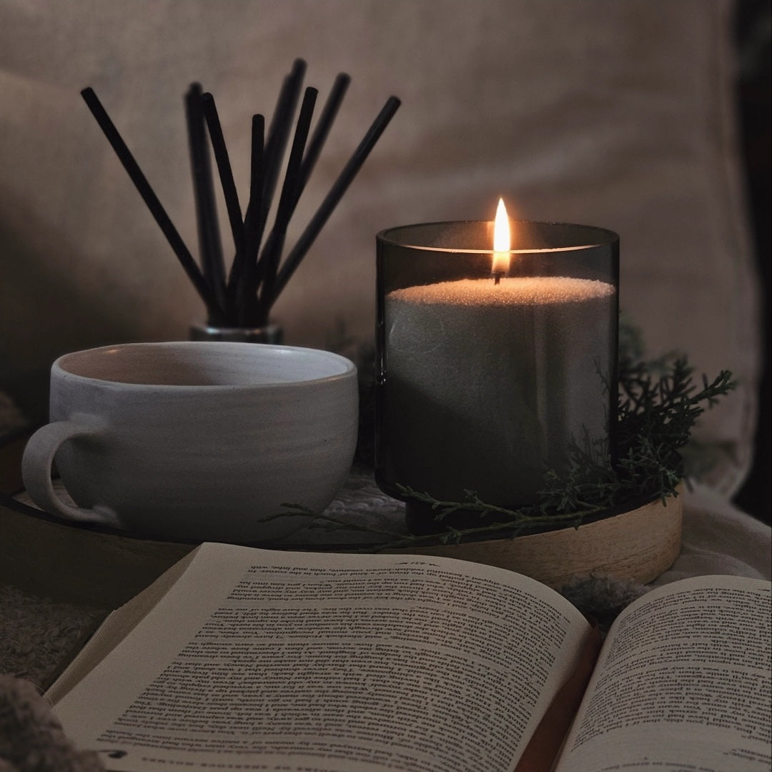 Smoky glass pearled candle beside an open book, coffee cup and reed diffuser in a cosy reading nook.