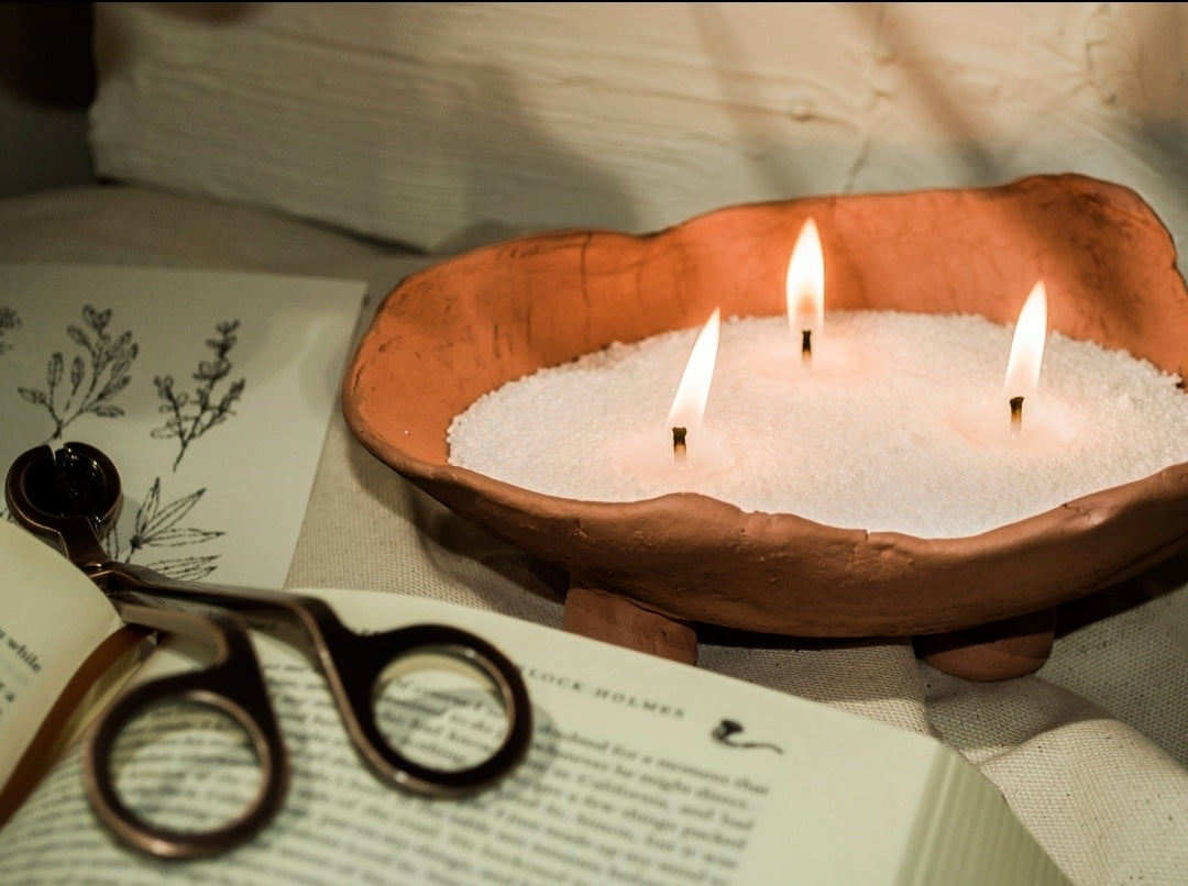 Three-wick white wax pearl candle in a rustic terracotta bowl beside an open book and reading glasses.