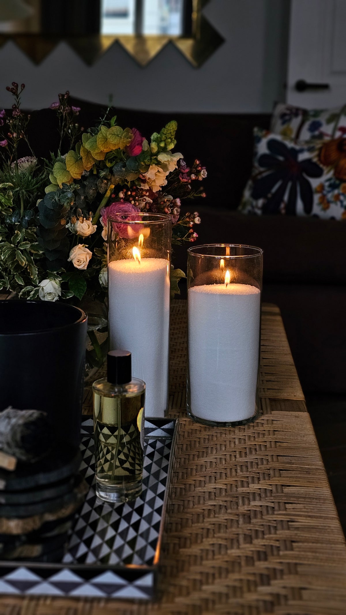 Two tall glass cylinder candles filled with white Hom33 wax pearls and three lit wicks, styled beside fresh flowers.
