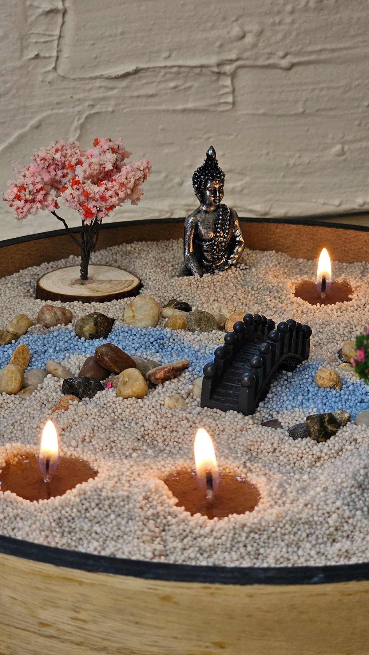 Zen garden candle sand display with a Buddha, bridge, stones, blossom tree and three lit wicks.